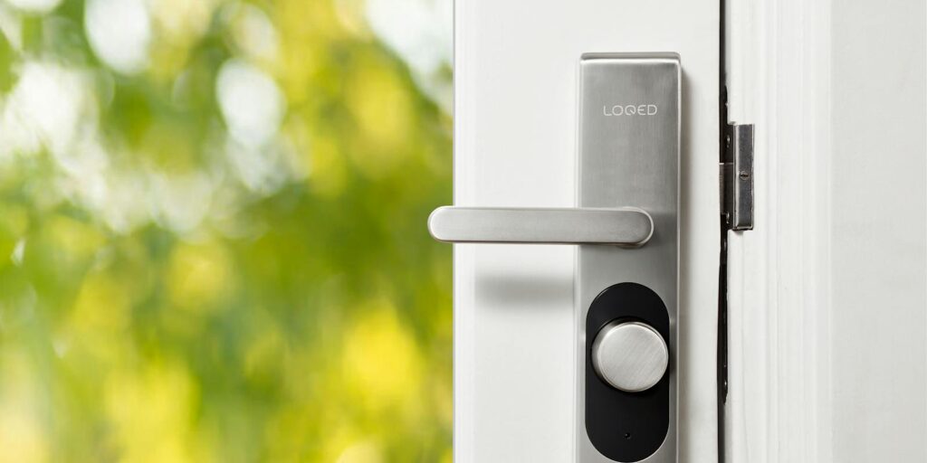 Sleek smart door lock on a white door with blurred green foliage in the background.